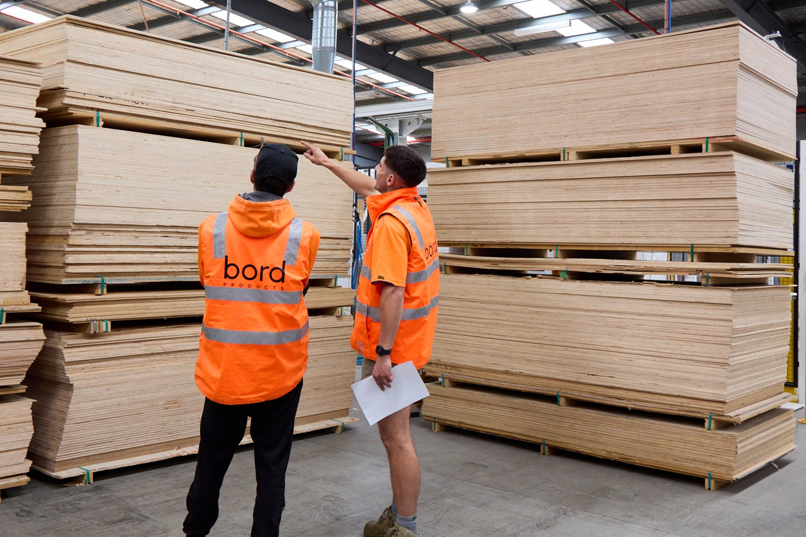 Two workers in orange vests inspecting stacks of wooden panels in a warehouse.
